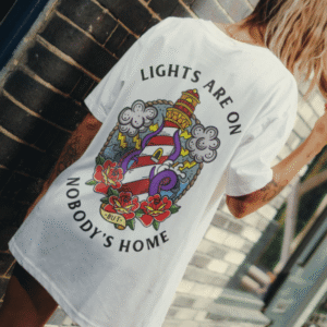 Person wearing the white Lights Are On But Nobody's Home graphic unisex T-shirt from Broken Society with a colourful lighthouse tattoo-style back print