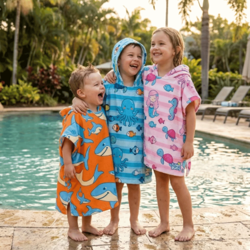 Children wearing FloatBuds Matching Poncho Towels in shark, blue sea life and pink mermaid designs beside a swimming pool